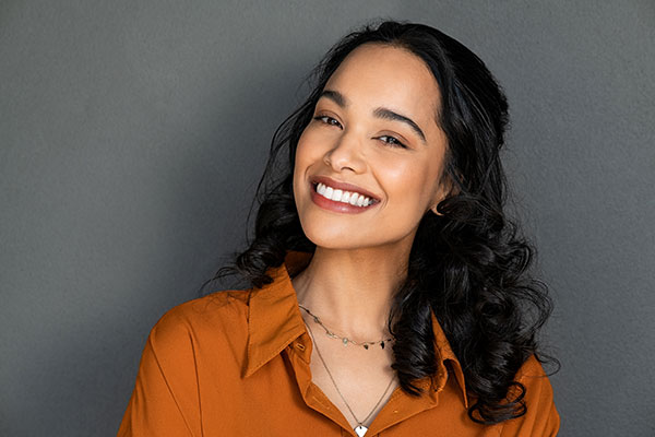 Smiling woman with dark hair, wearing a peach blouse and a necklace, posing against a neutral background.