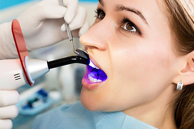 The image shows a woman in a dental chair receiving dental treatment, with a dentist using a device to clean or inspect her teeth.