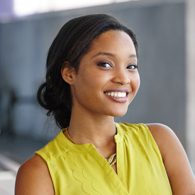 A smiling woman with dark hair, wearing a yellow top and posing against a blurred background.