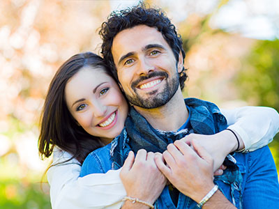 A man and a woman, possibly a couple, are smiling at the camera. The man is wearing a short beard and has his arm around the woman s shoulder. They appear to be standing in front of a window with blinds partially drawn.