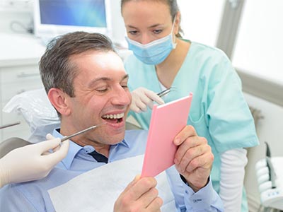 A man seated in a dental chair, holding up a pink card with a smile, while a female dentist looks on.