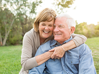 The image is a photograph of an older man and woman, both smiling, embracing each other in a park setting during daylight. They are standing on grass with trees in the background.