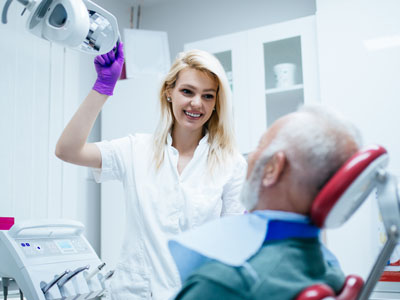 The image depicts a dental professional, likely a dentist or dental hygienist, assisting an elderly patient in a dental chair.