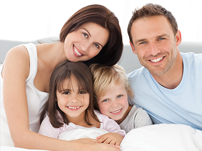 This is a color photograph featuring a family of four adults and two young children, all smiling and posing together in what appears to be a bedroom setting.