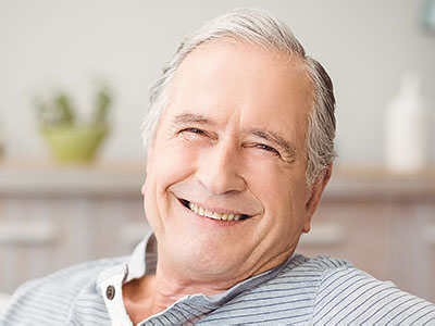 The image shows an elderly man with gray hair, smiling and looking directly at the camera. He appears to be seated comfortably in a home setting.