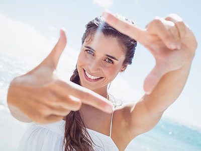 A young woman with a radiant smile is holding up her index finger, creating a lens effect in the foreground of the image. She appears to be at a beach or seaside location, as suggested by the bright daylight and the water visible in the background.